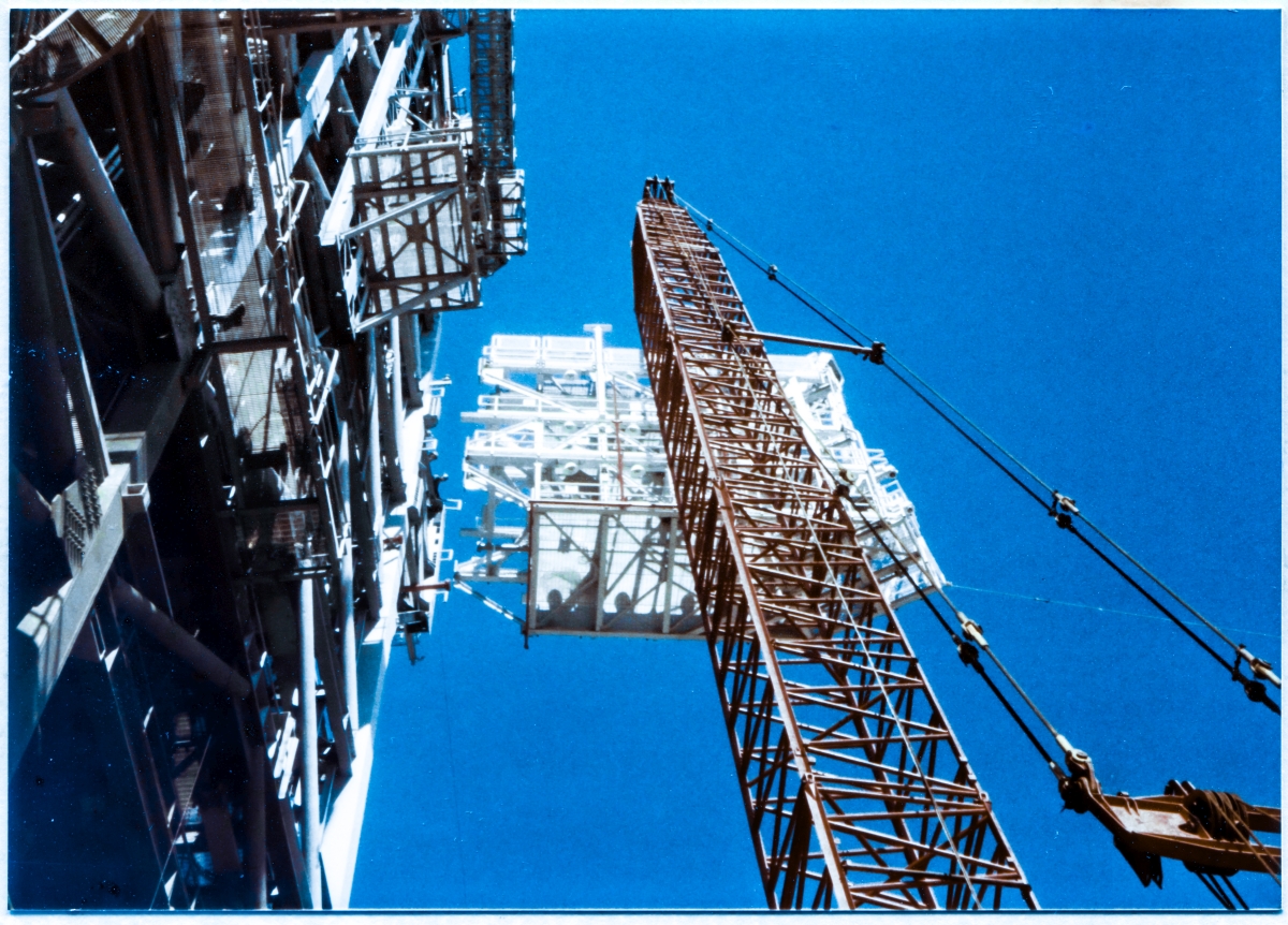 Image 124. One hundred thousand pounds of IAA continues to close in on direct contact with its Support Structure on the FSS, over one hundred fifty feet above where you're seeing it from the Pad Deck at Space Shuttle Launch Complex 39-B, Kennedy Space Center, Florida. A mass like that, even when moving at a snail's pace, carries a TREMENDOUS inertia, and will very much want to keep going right on through things, violently bashing them out of its path with a fearsome and uncanny slow motion way about it, as opposed to simply coming to a halt and resting against them. This is an extremely dangerous time and situation. Alignment is crucial, and the unseen Union Ironworker from Local 808 working for Ivey Steel, far out of frame to the right, down on the Pad Deck, holding the tag line which controls the rotation of the IAA, needs to be positioned precisely to pull it into the one and only correct orientation. He is being given instructions to do so by the pair of ironworkers that you can see leaning out over the top of the precipice, standing on the Support Stucture at elevation 220'-0”, carefully monitoring the situation and giving what amounts to life-and-death instructions to both their fellow ironworker and the crane operator, who cannot even see what's happening in the narrowing spaces between the structural steel elements of the IAA and the FSS which it will soon be in hard contact with. The moment of truth is rapidly approaching, and it simply cannot be gotten wrong. Photo by James MacLaren.